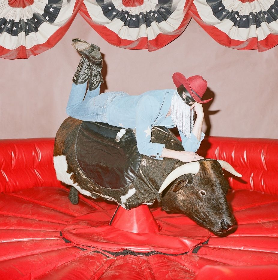 Person in a denim outfit, cowboy hat and mask riding a mechanical bull with patriotic bunting behind them.