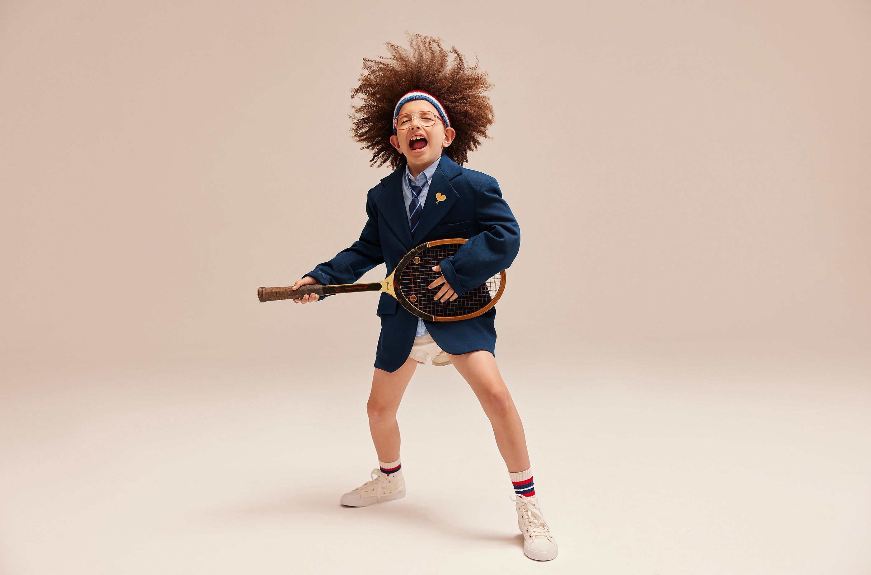 Child in a blue blazer and striped headband holding a tennis racket, mid-shout in a studio portrait.