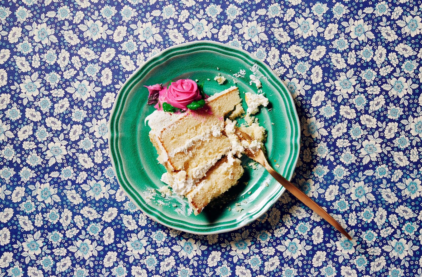 A slice of layered cake with white frosting and a pink frosting rose, on a green plate with a fork.