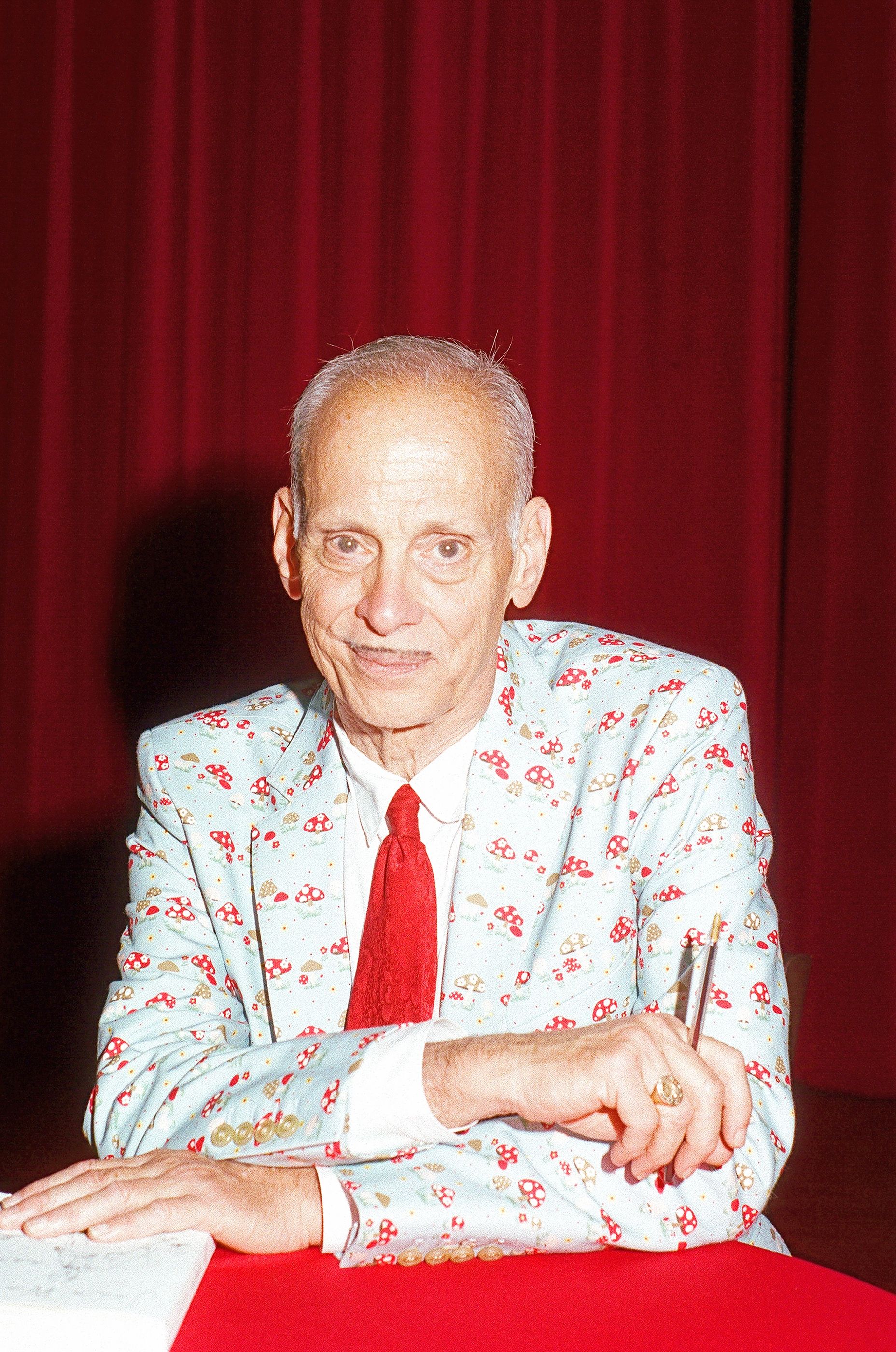 Portrait of an older man in a mushroom-print suit and red tie, seated at a table against a red curtain.