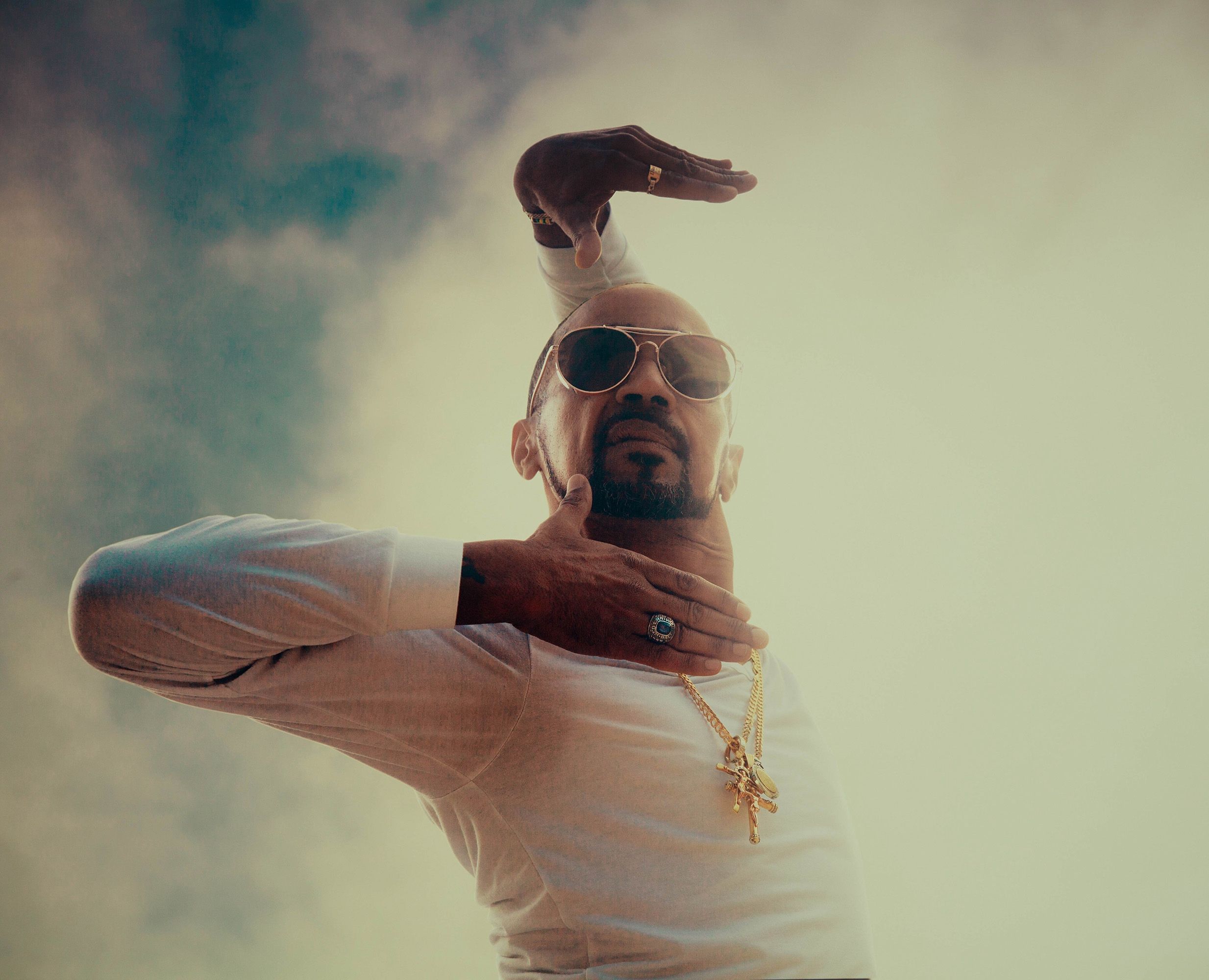 Man making a dabbing gesture, wearing sunglasses and gold chains, smoky beige background.