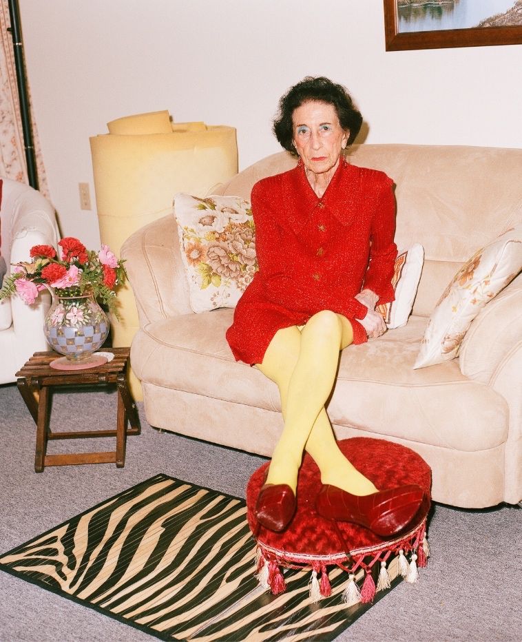 Elderly woman in a red outfit and yellow tights sitting on a cream sofa in a living room.