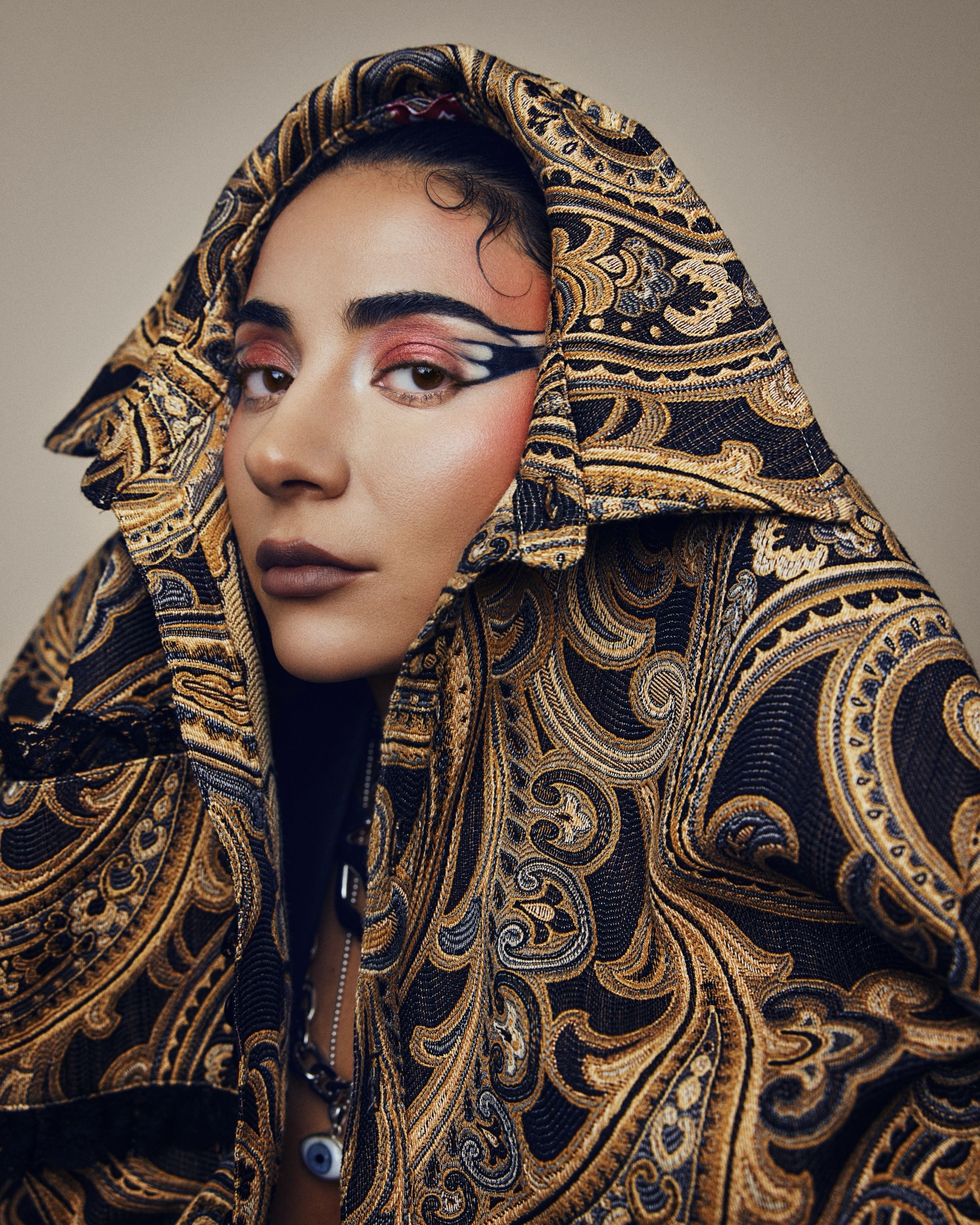 Woman is draped in a gold and black paisley brocade fabric worn as a hood, with dramatic red and black eye makeup.