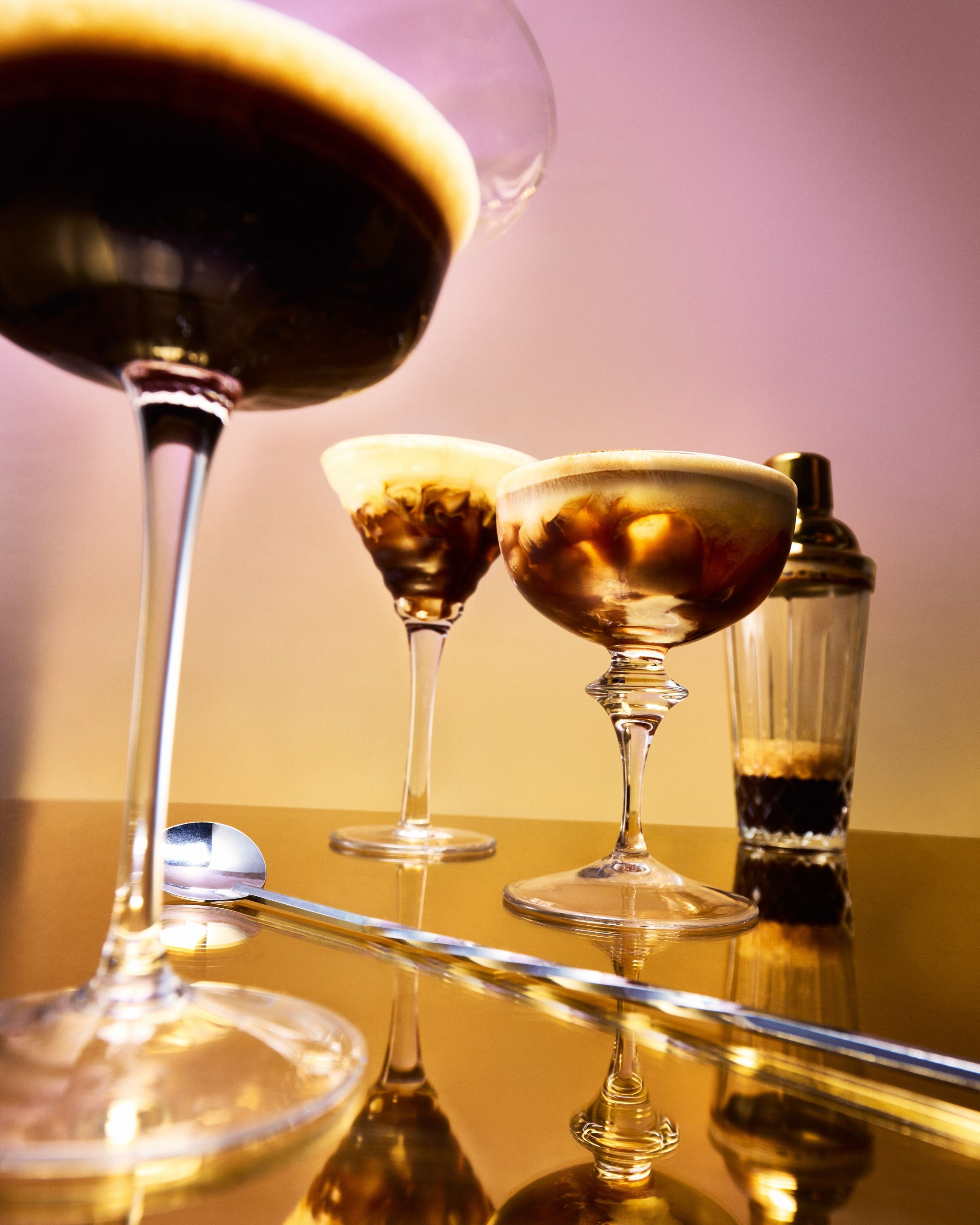 Several espresso martinis in coupe and martini glasses on a mirrored surface, with a cocktail shaker.
