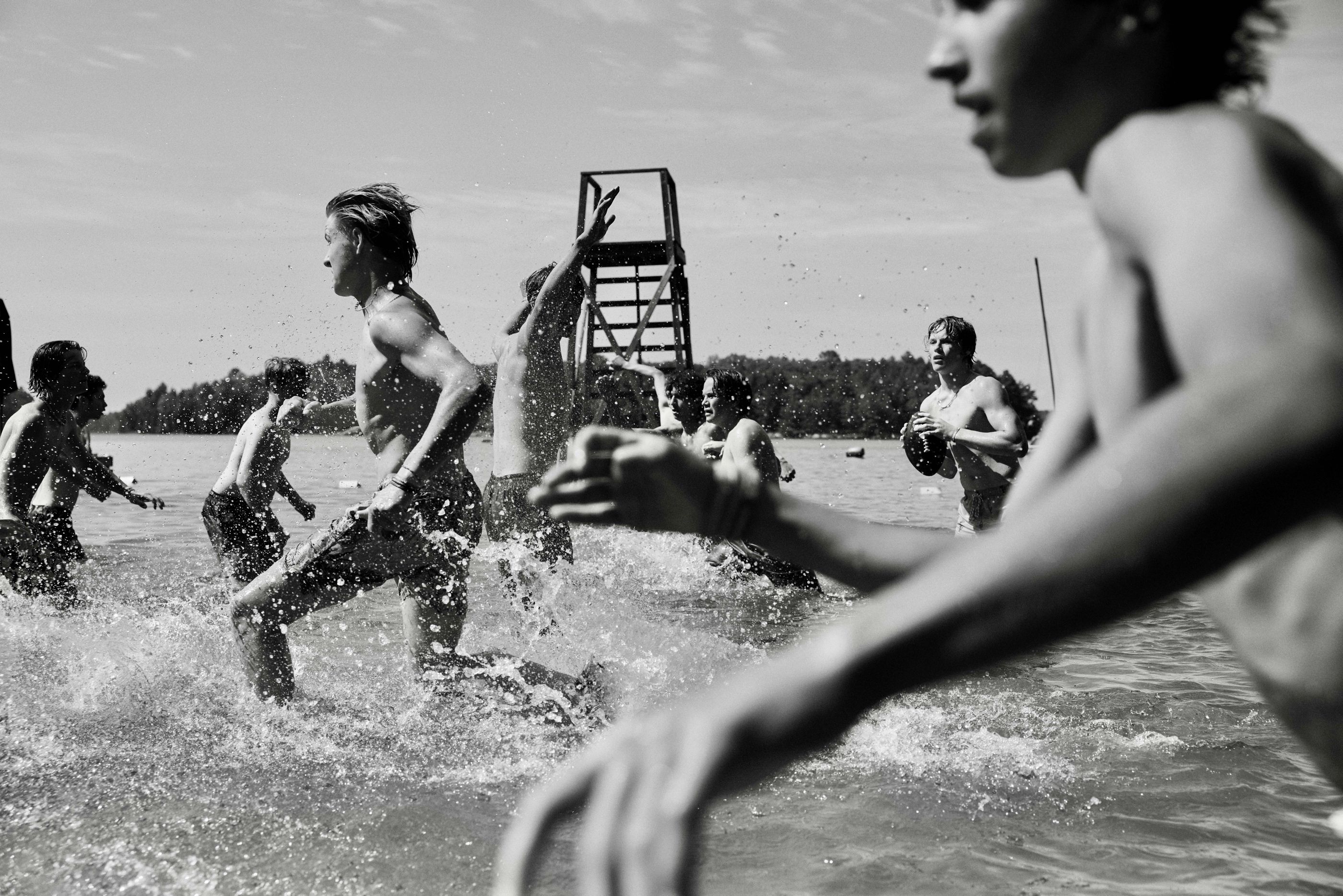 Black-and-white photo of teen boys playing football in lake water, splashing as they run.