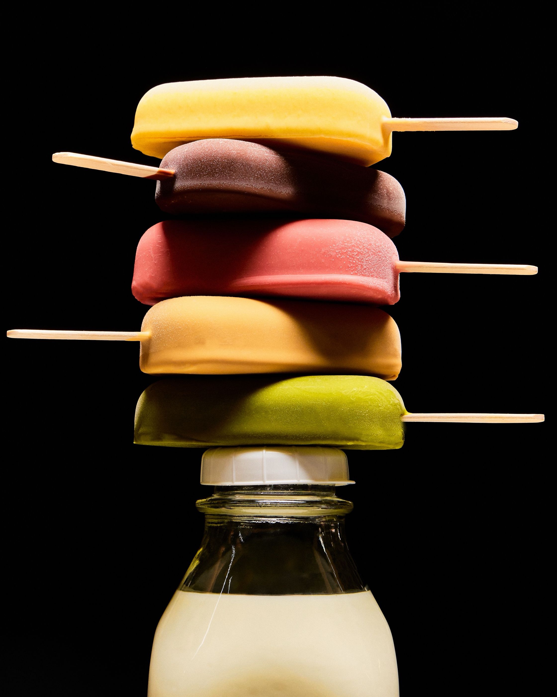 Five different colored popsicles on sticks stacked on top of a glass milk bottle, on black.