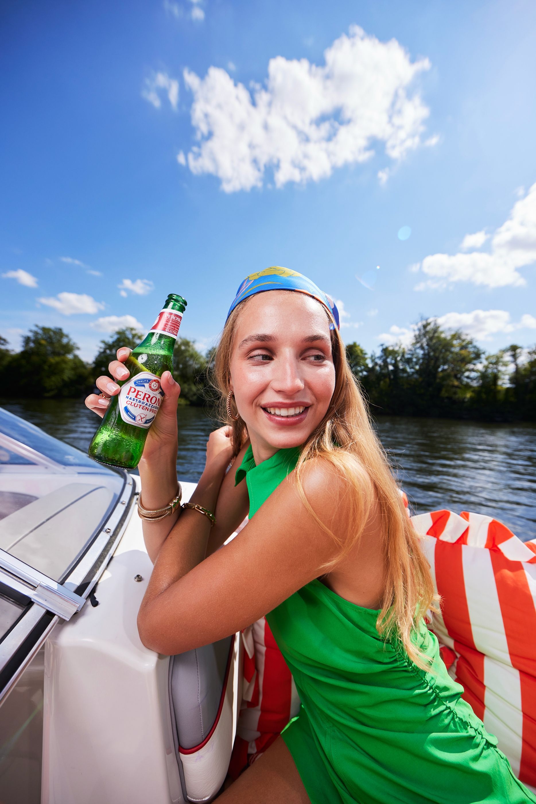 Smiling woman in a green top and blue headscarf holds a Peroni bottle on a boat, with a river and trees behind her.