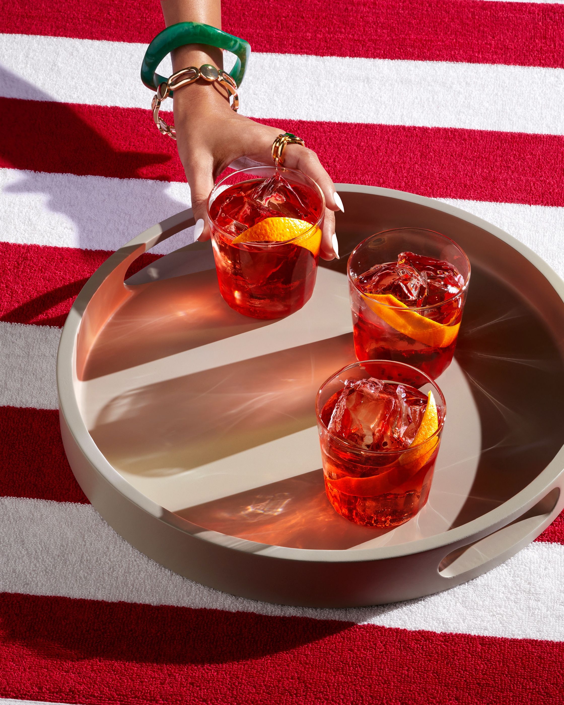 A hand reaches for one of three red cocktails with orange peel on a round tray, set on a red and white striped towel.