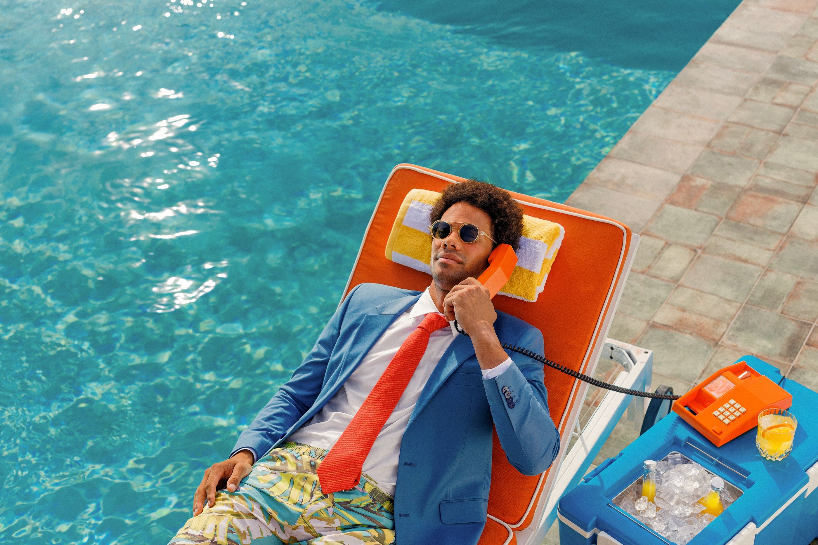 Man in a blue suit and tie over swim shorts lies on a pool lounger, talking on an orange corded phone.