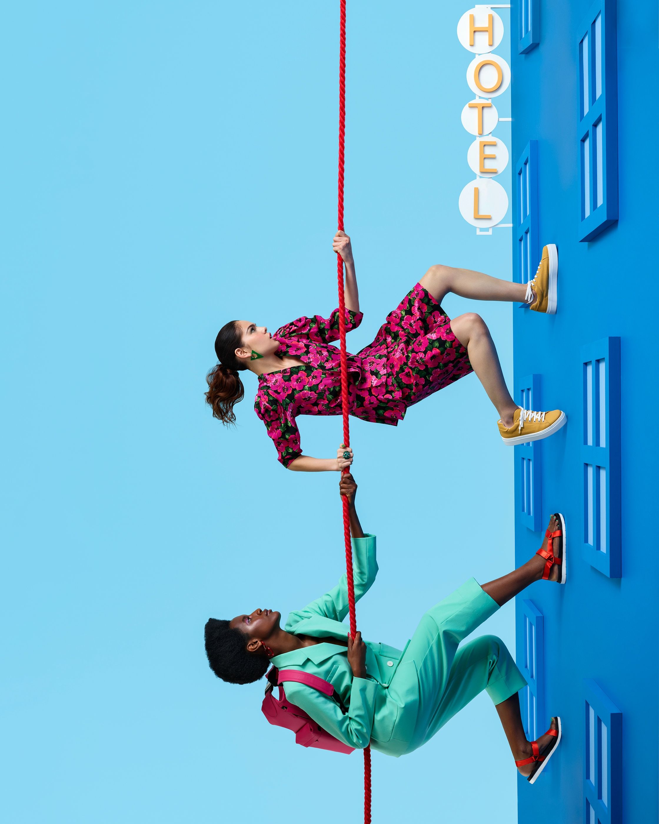 Two people in a floral dress and a mint suit grip a red rope, feet on a blue wall below a HOTEL sign.