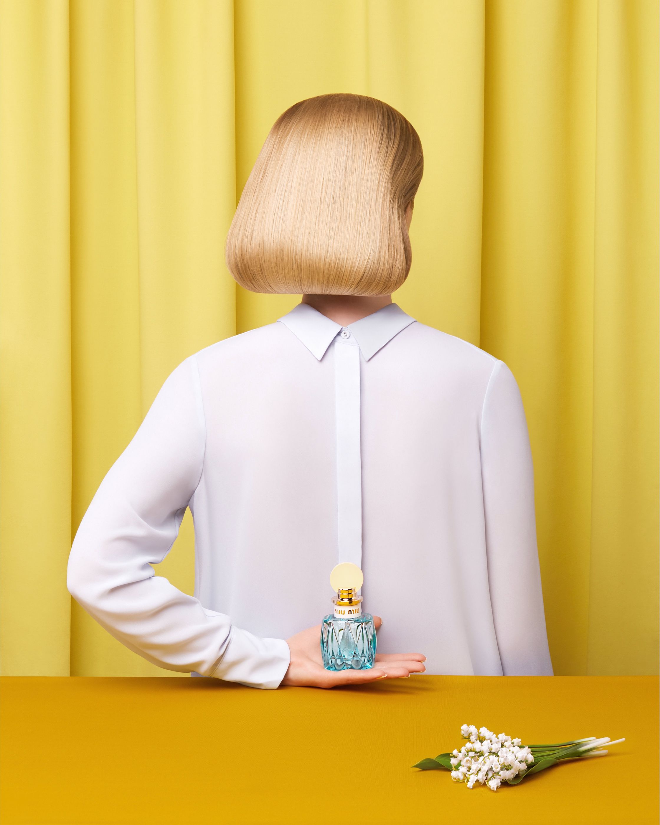 Woman seen from behind, her head hidden by a blunt blonde bob, holds a Miu Miu perfume bottle behind her back.