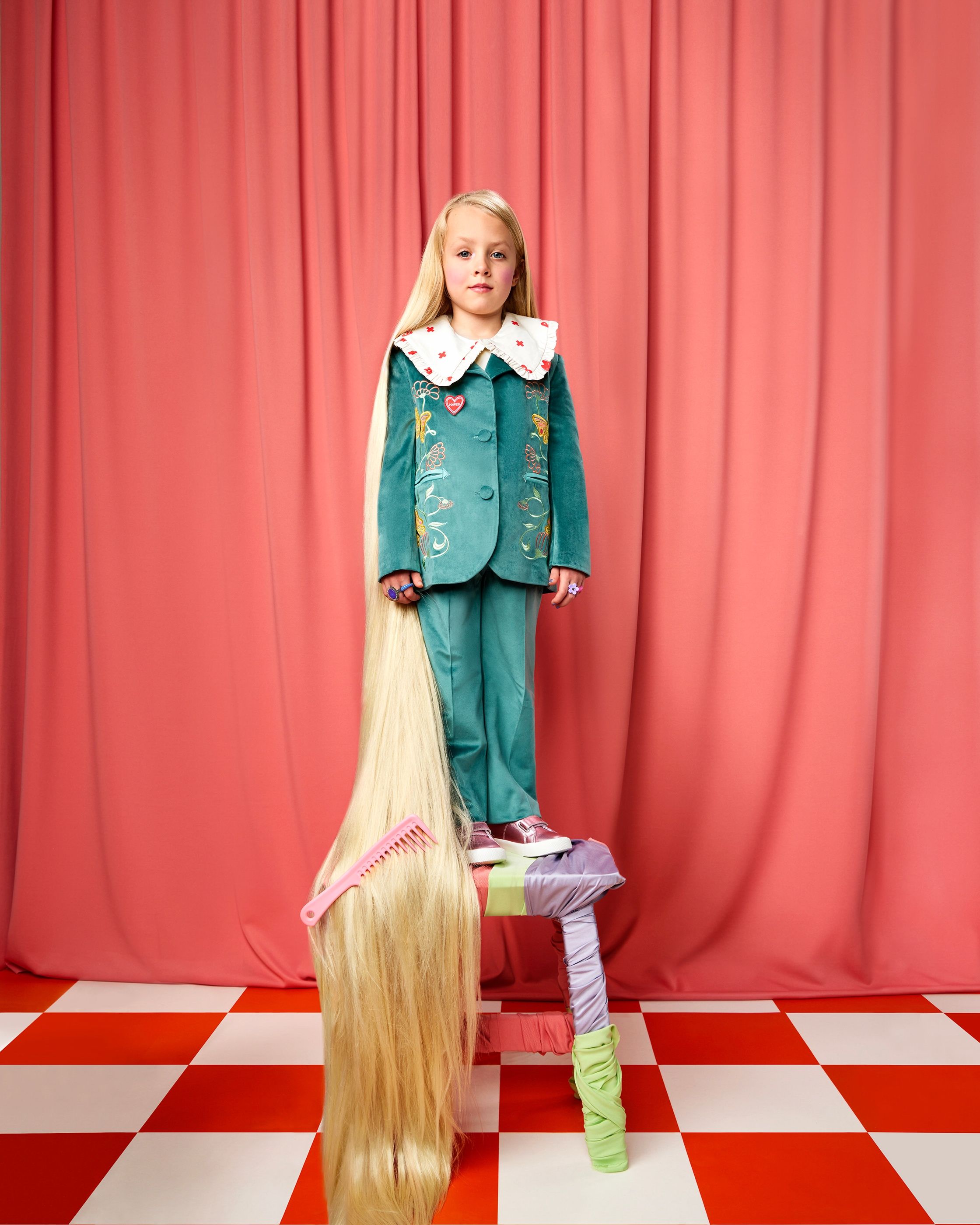 Young girl in a teal velvet suit stands on a stool, her blonde hair reaching the floor, in front of a pink curtain.