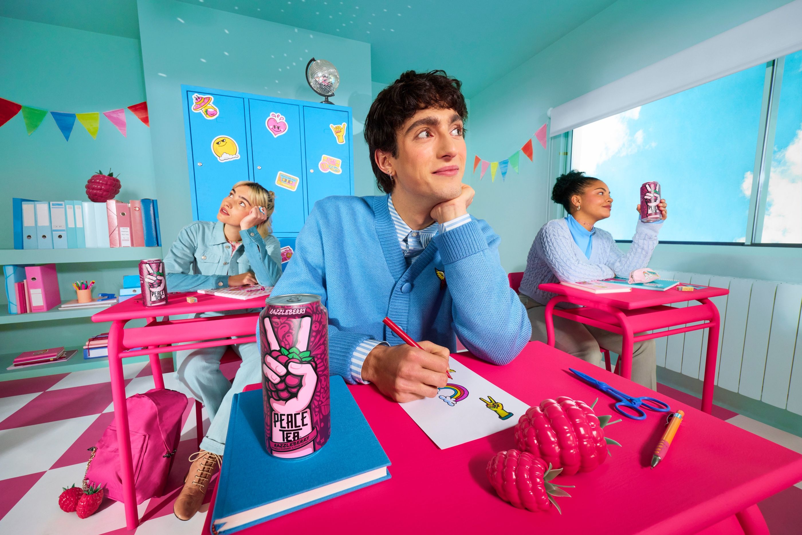 Student in a blue cardigan draws at a pink desk with a Peace Tea can, in a teal classroom with two classmates.