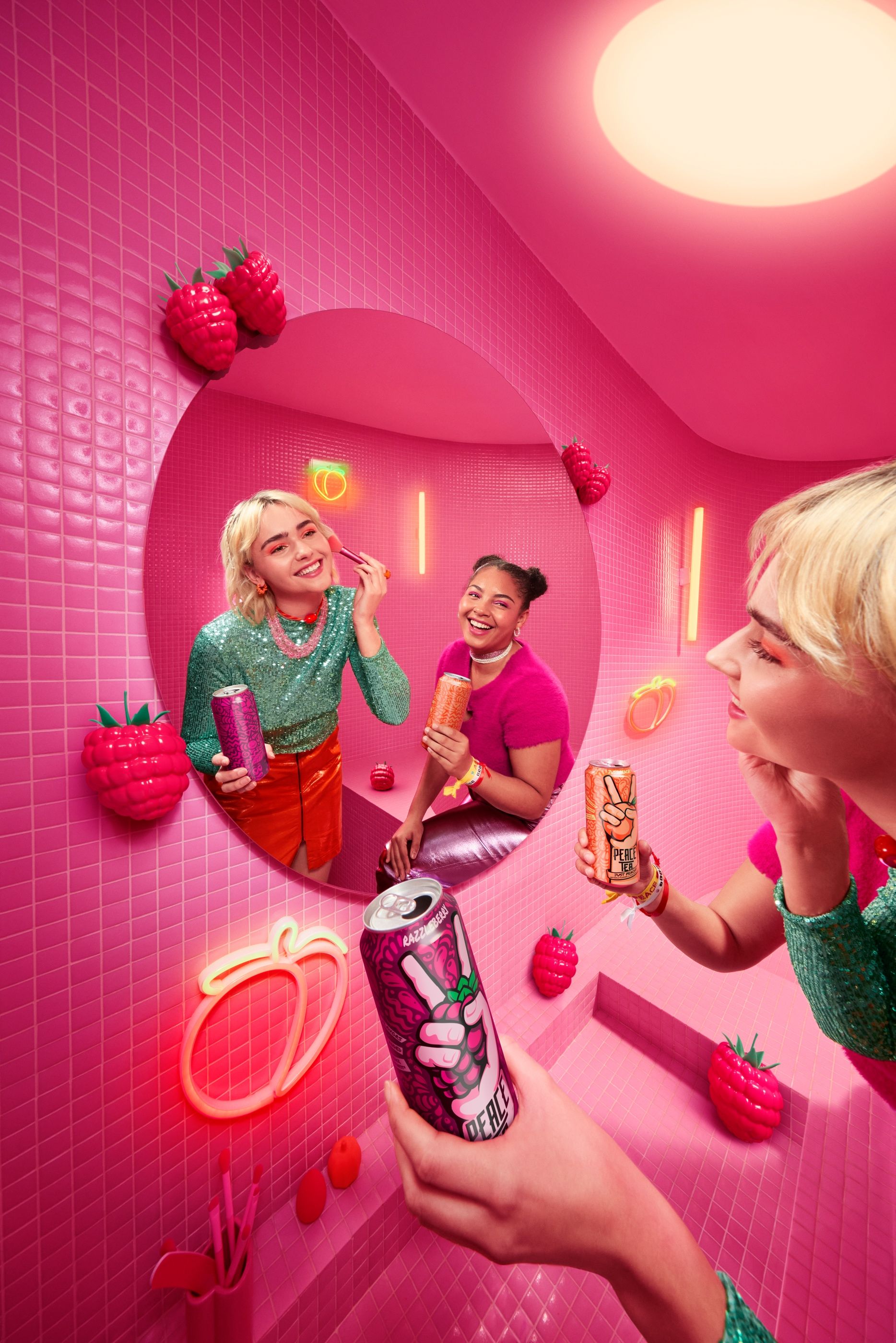 Two women in a pink tiled bathroom hold Peace Tea cans, one applying makeup at a round mirror.