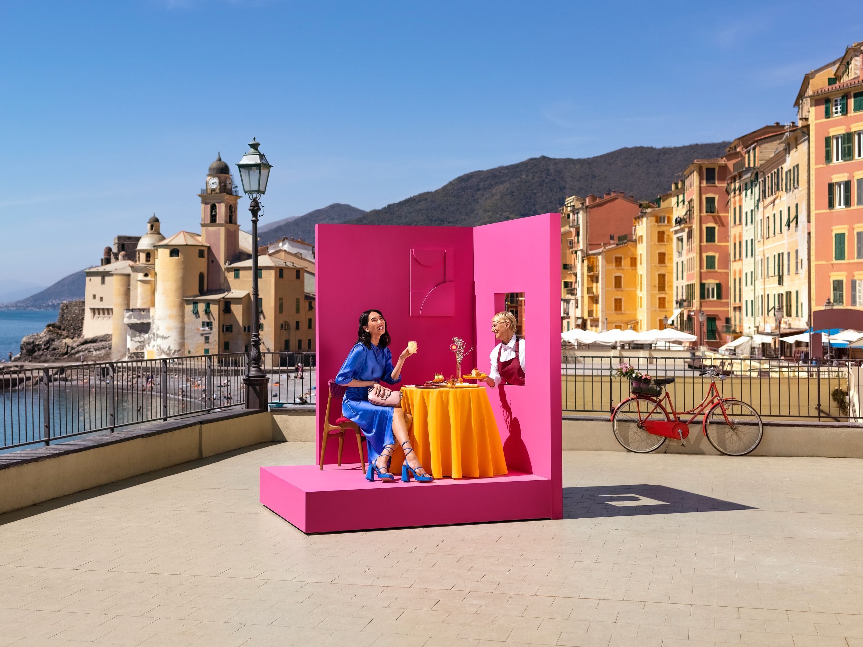 Woman in a blue dress laughs at a set table inside a pink stage set on a seaside promenade as a server passes a dish.