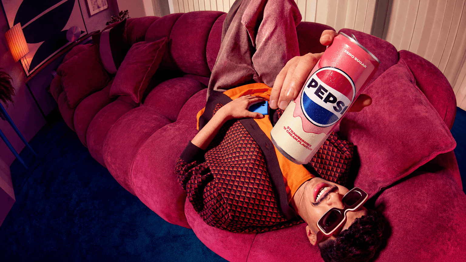 Person in sunglasses reclines on a plum sofa holding a pink Pepsi can