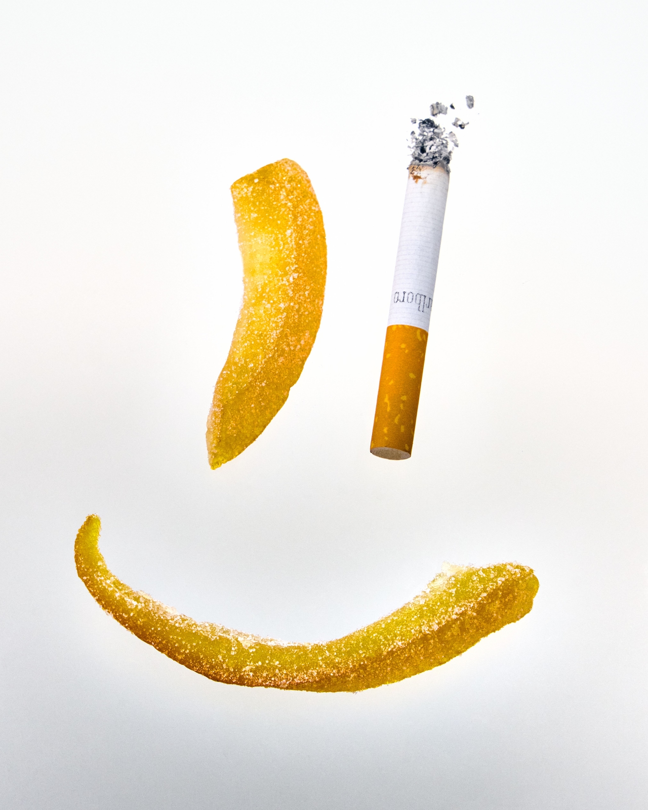 A still-life image of a lit cigarette with falling ash, positioned between two curved strips of golden-yellow citrus peel  against a pale background.