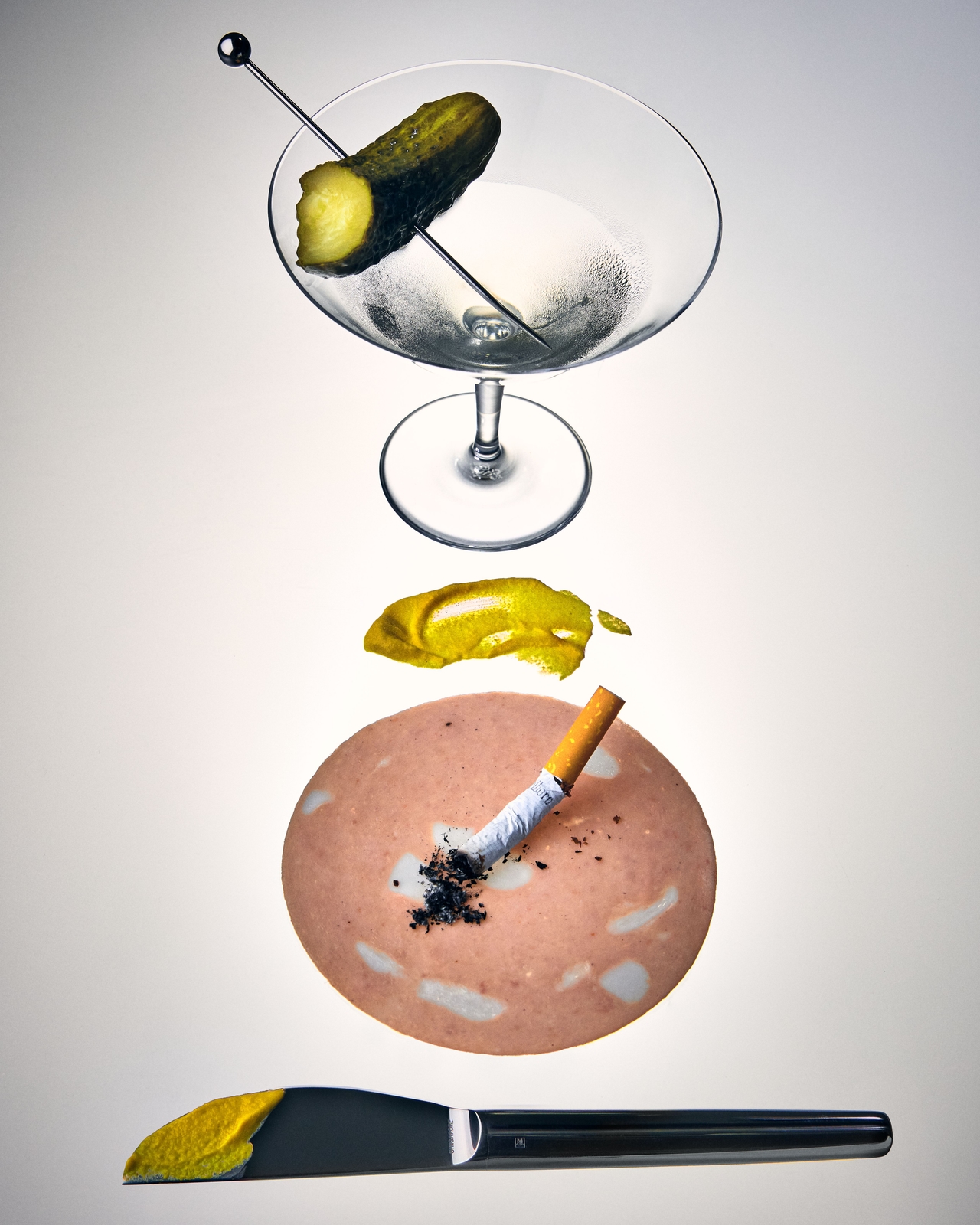 A still-life composition featuring a martini glass garnished with a pickle, a cigarette on a piece of deli meat, and a black knife with a smear of mustard on the blade, arranged vertically against a pale background.