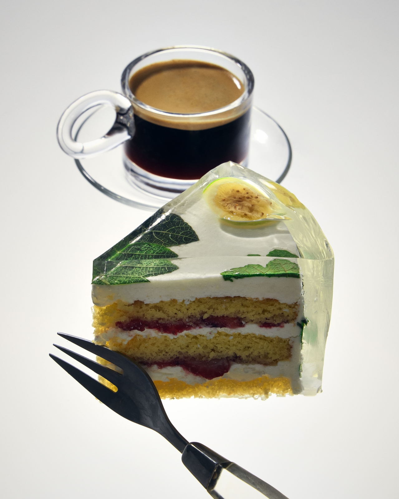 A slice of layered sponge cake with cream, red berry filling, green leaves, and a slice of passion fruit in clear gelatin, served with a cup of espresso on a white surface. A black dessert fork rests beside the cake.