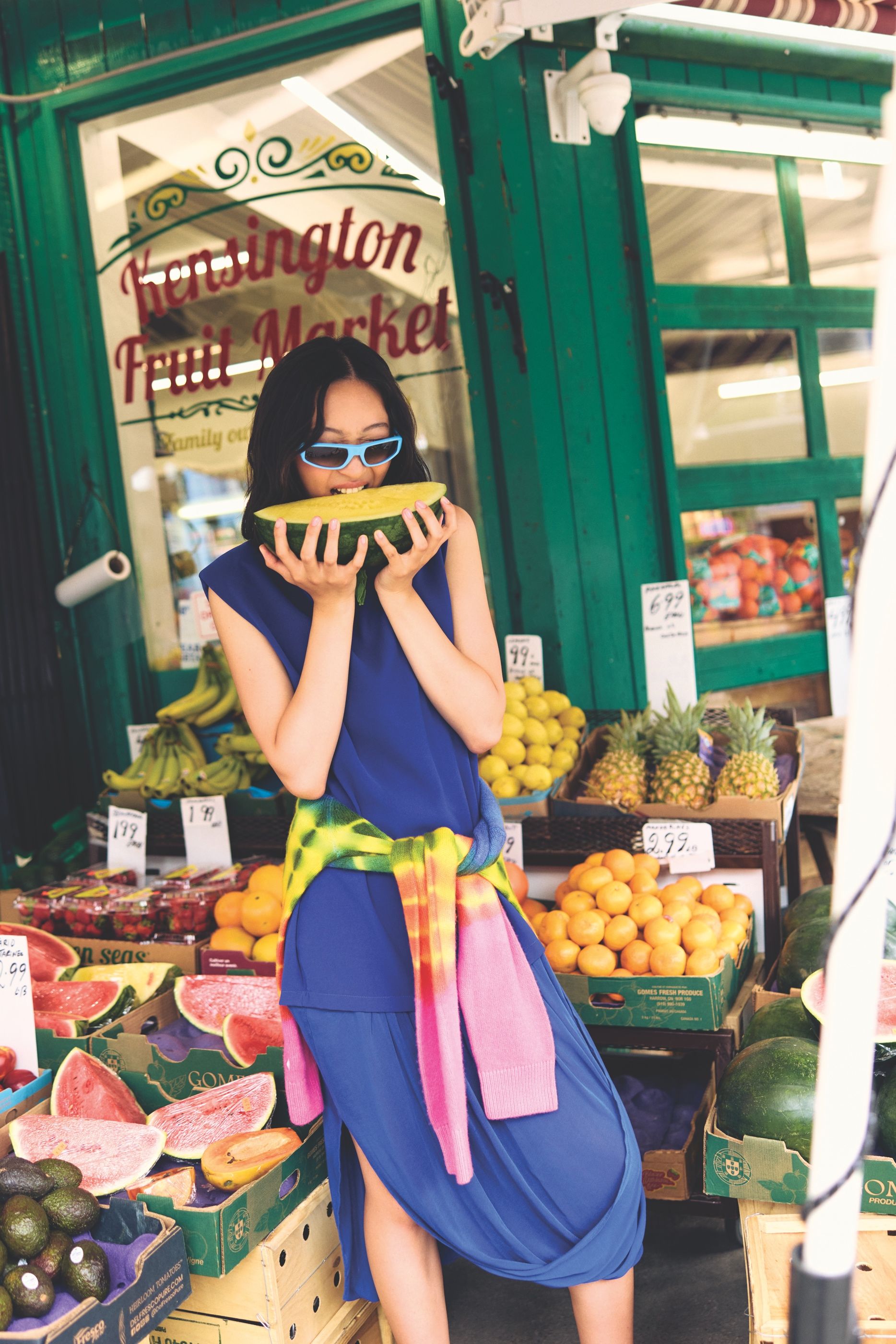 A woman wearing a cobalt blue dress, blue sunglasses, and a colourful sweater tied around her waist holds a slice of watermelon near her face at an outdoor fruit market surrounded by fruit