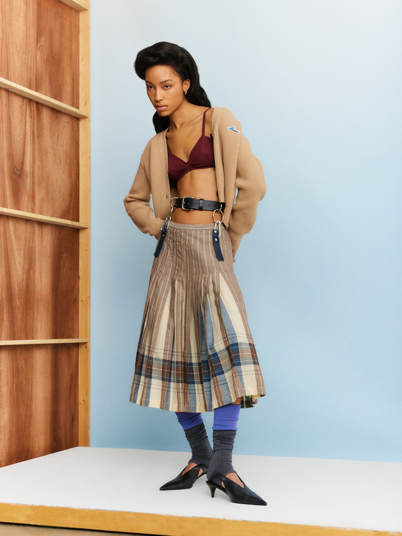 A model poses in a burgundy bralette, beige knit cardigan, pleated plaid midi skirt, black belt with suspenders, blue leg warmers, grey socks, and black pointed-toe heels against a light blue studio backdrop with wooden shelving