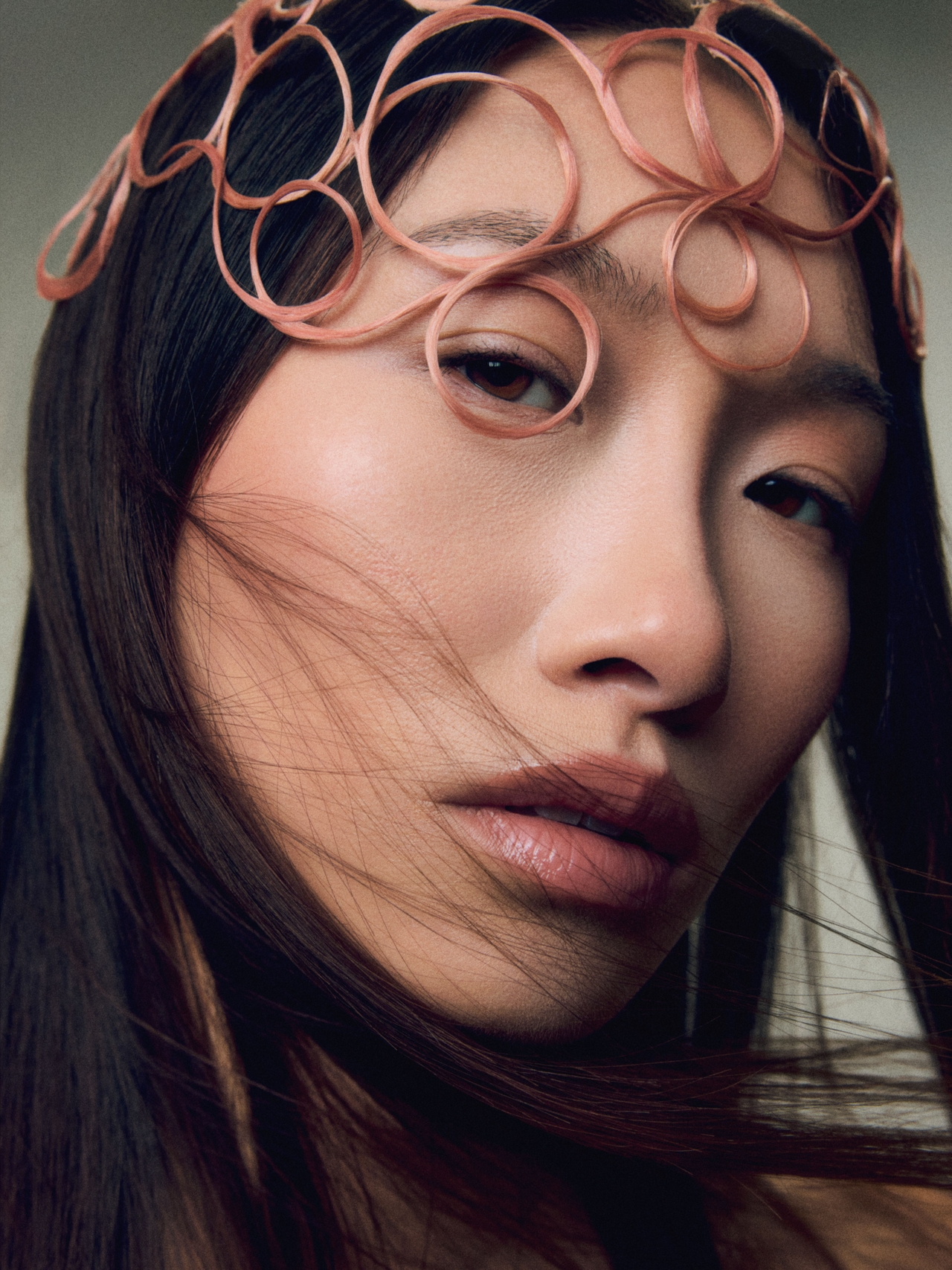 Close-up portrait of a model with long, dark hair, wearing intricate rose-pink looped wire accessories across her forehead. Loose strands of hair frame her face against a grey background.